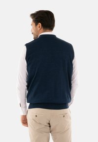 Gilet lavorato a maglia blu navy con orlo a coste. Indossato sopra una camicia bianca a maniche lunghe. Design semplice, senza motivi o accenti visibili.
