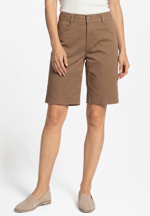 Frau trägt knielange braune Shorts, weißes ärmelloses Oberteil und beige Slip-On-Schuhe, steht vor einem schlichten weißen Hintergrund.