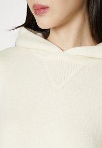 Femme portant un sweat à capuche en maille de couleur crème, montrant l'encolure et le bas du visage avec des lèvres naturellement brillantes et des cheveux raides et foncés.