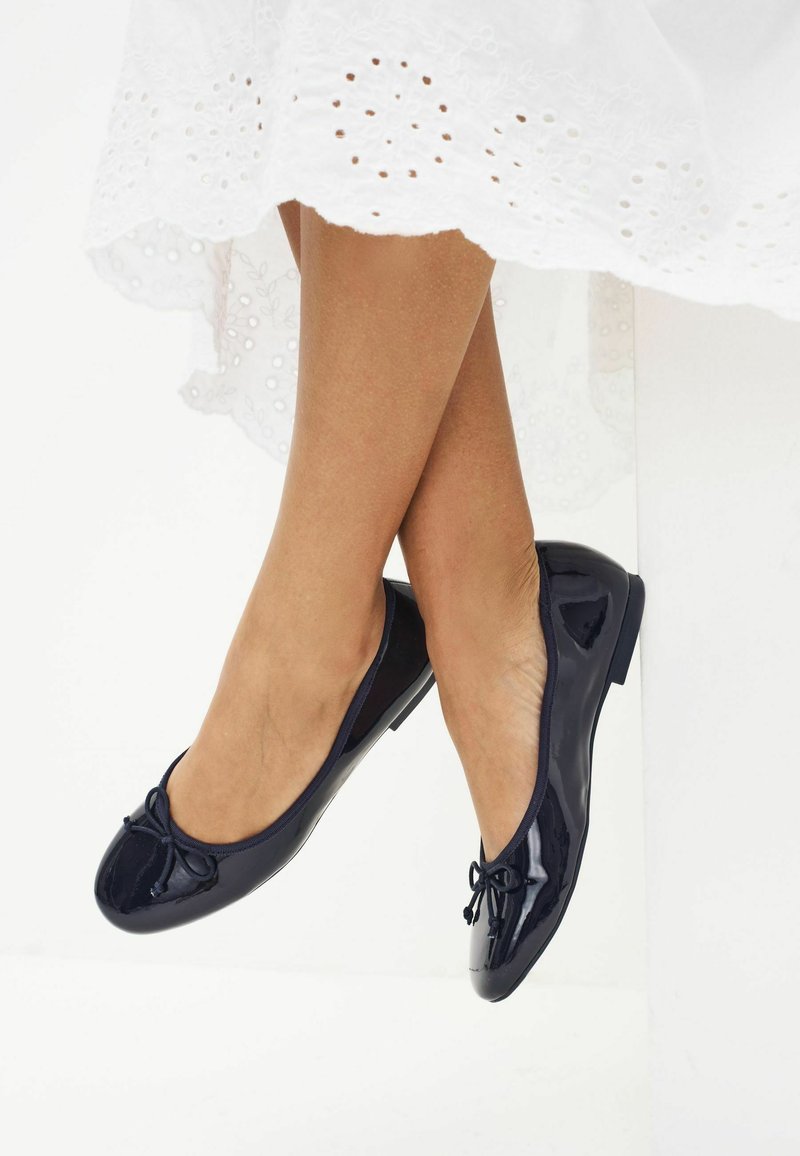 Marineblaue Patent-Ballerinas mit runder Zehenpartie, dezentem Schleifendetail und niedrigem Absatz, vor dem weißen bestickten Saum eines Kleides.