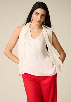 Donna con capelli lunghi neri indossa una camicetta bianca senza maniche e pantaloni rossi, posando con le mani sui fianchi davanti a uno sfondo neutro.