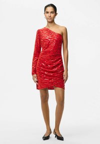 Raudona blizgučiais suknelė su viena ilga rankove, asimetrišku iškirpimu, raukšlėta konstrukcija ir trumpa ilgio, derinama su juodais smailiais aukštakulniais.