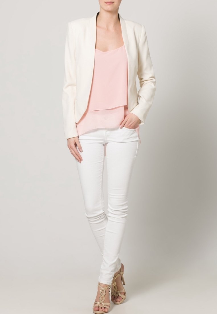 Femme portant un blazer blanc, un chemisier rose pâle, un pantalon skinny blanc et des sandales à talons beiges, debout devant un fond uni.
