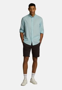 Jasnoniebieska koszula z długim rękawem, podwiniętymi rękawami, ciemne szorty i białe sneakersy. Koszula ma klasyczny kołnierzyk i luźny krój.