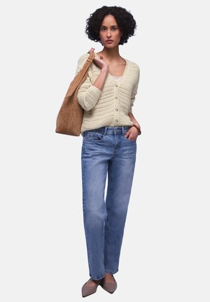 Donna con capelli ricci che indossa un cardigan beige all'uncinetto, jeans blu, ballerine marroni e porta una grande borsa a tracolla in suede marrone.