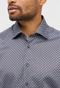 Camicia con colletto caratterizzata da un motivo geometrico in blu scuro, nero e bianco. Parte anteriore con bottoni e una texture liscia.