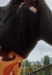 Sudadera de lana negra con media cremallera y bolsillo delantero, con un logo multicolor. Leggings estampados con formas abstractas cálidas.