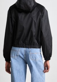 Svart, lättvikts, zip-up jacka med huva, med en strukturig yta och elastisk midja. Kombinerad med ljusblå raka jeans.