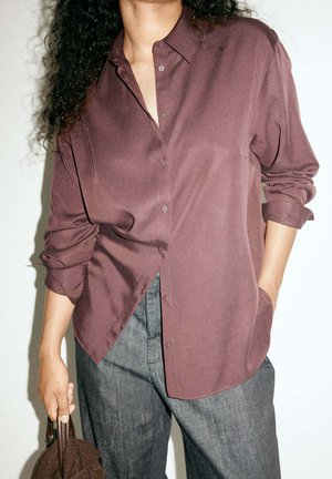 Persona con cabello rizado lleva una camisa burdeos suelta con botones y pantalones grises, sosteniendo una pequeña bolsa marrón texturizada en una mano.