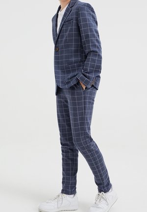 Granatowy garnitur w kratę o dopasowanym kroju, z pojedynczym guzikiem, klapami z wycięciem oraz dopasowanymi spodniami, zestawiony z białymi trampkami.