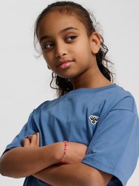 Jong meisje met krullend haar, draagt een blauw shirt met een klein bij-logo, armen gekruist, kijkt naar de zijkant met een lichte glimlach.