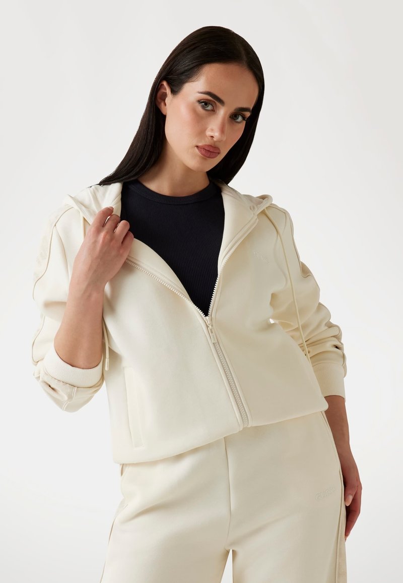 Felpa zip con cappuccio in crema, con cappuccio regolabile, polsini a costine e vestibilità rilassata, abbinata a pantaloni crema coordinati con dettaglio del logo.