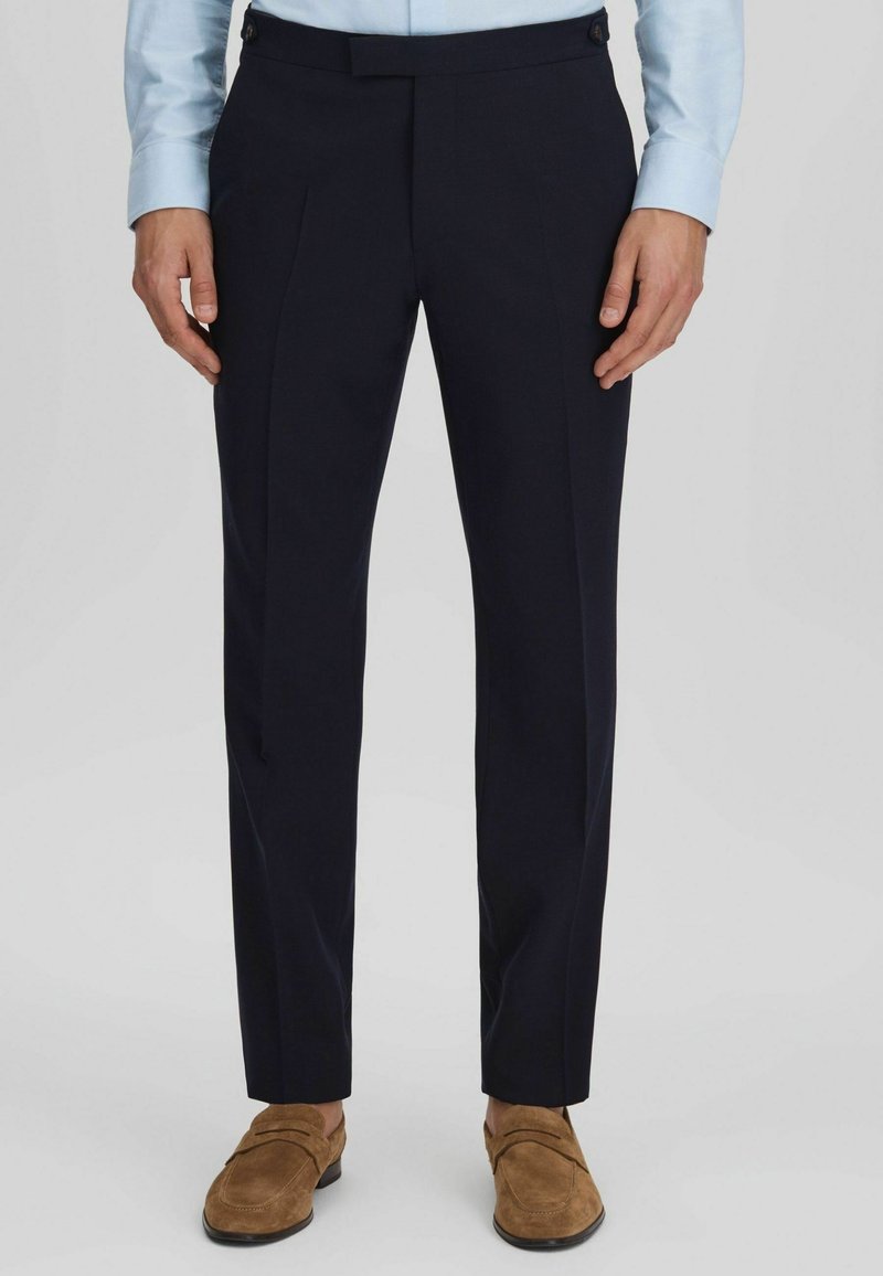 Homme portant un pantalon de costume bleu marine foncé sur mesure avec des mocassins en daim beige et une chemise bleu clair, debout devant un fond uni.