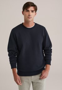 Hanorac bleumarin dintr-un material texturat, cu guler rotund și manșete cu model de nervuri. Croiala este lejeră, cu o margine dreaptă și fără aplicații.
