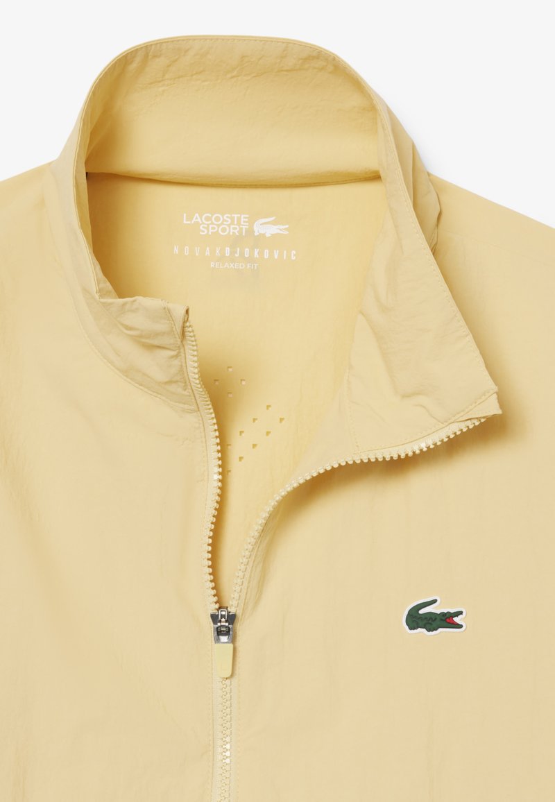 Beige Lacoste Sport jas met halve rits, opstaande kraag, Novak Djokovic label aan de binnenkant en geborduurd groen krokodillenlogo op de borst.