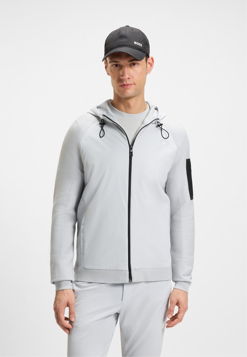 Egy férfi világosszürke cipzáras kapucnis felsőben és hozzá illő nadrágban áll, fekete "BOSS" feliratú sapkát viselve, egy sima fehér háttér előtt.