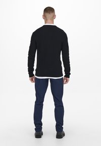 Pull en maille noir à manches longues et poignets côtelés, associé à un pantalon bleu marine. L'ensemble comprend un sous-vêtement blanc en couches.