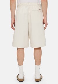 Henkilö, jolla on päällään polvenmittaiset beigehousut, joissa on joustava vyötärö, valkoiset sukat ja sandaalit. Näkymä on takaa.