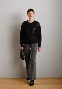 Černý svetr s flitrovými detaily, šedé džíny a černé kotníkové boty. Modelka drží malou černou kabelku na neutrálním pozadí.