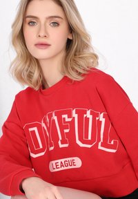 Piros kapucnis pulóver, amelynek túlnőtt fehér felirata van: "OYFUL LEAGUE". Kerek nyakkivágással és bordázott mandzsettával készült puha pamut anyagból.