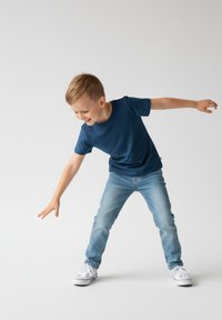 Granatowy t-shirt z krótkim rękawem, okrągły dekolt, dżinsy i białe trampki. Dziecko w zabawnej pozie na neutralnym tle.