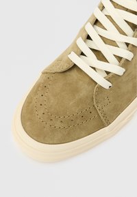 Καφέ σουέντ αθλητικά παπούτσια με κρεμ κορδόνια, στρογγυλή μύτη, λεπτομέρειες από διάτρητο σχέδιο και υφή καουτσούκ στην σόλα. Κοντινή λήψη.