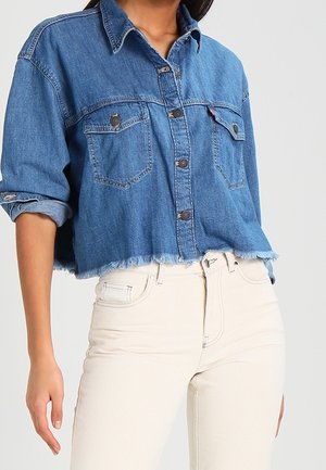 Woman wearing a blue cropped denim jacket with rolled-up sleeves and high-waisted off-white pants against a plain background.