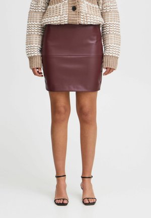Gonna in finta pelle bordeaux con una forma aderente, texture liscia e dettagli delle cuciture visibili, indossata con décolleté nere con cinturino alla caviglia.