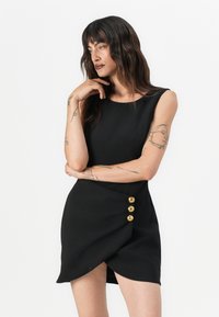 Robe noire sans manches avec un col arrondi, design enveloppant et trois boutons dorés sur le côté. Tissu lisse sans motifs visibles.