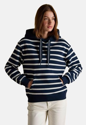 Young woman with medium-length brown hair wearing a navy blue and white striped hoodie and white pants, hands in hoodie pocket.