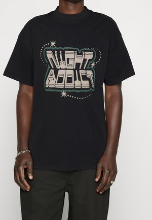 Czarny, oversize'owy t-shirt z beżowym, retro blokowym napisem "NIGHT ADDICT" otoczonym wzorami gwiazdek i kropek, noszony z ciemnymi spodniami, srebrną bransoletką i pierścionkiem.