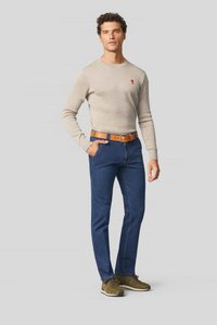 Beige knitted sweater with a small red emblem, blue jeans, and a brown leather belt. Brown shoes with a textured finish complete the outfit.
