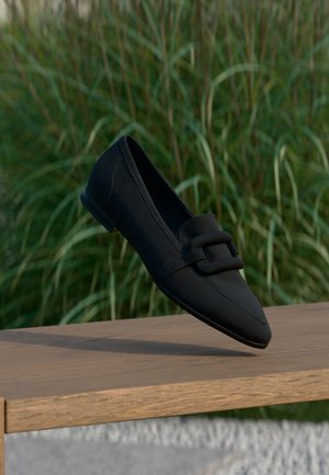 Zwarte loafer met puntige neus en decoratieve gesp, rustend op een houten oppervlak tegen een achtergrond van hoog groen gras.