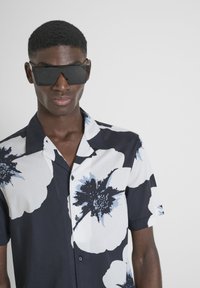 Camicia a maniche corte con stampa floreale nera, bianca e blu. Presenta un colletto e sei bottoni. Il modello indossa occhiali da sole neri.