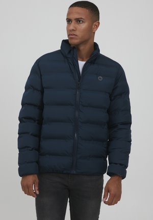 Chaqueta acolchada azul marino, diseño acolchado, cuello alto, cremallera frontal y detalle del logo en el pecho. Combinada con una camisa blanca y jeans oscuros.