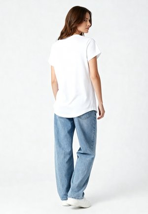 Vrouw in wit shirt met korte mouwen, losse blauwe spijkerbroek en witte sneakers, staand, iets gedraaid met handen in de zakken, tegen een effen achtergrond.