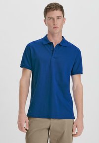 Blue polo shirt made of textured fabric, featuring a collar and three-button placket, paired with khaki pants.