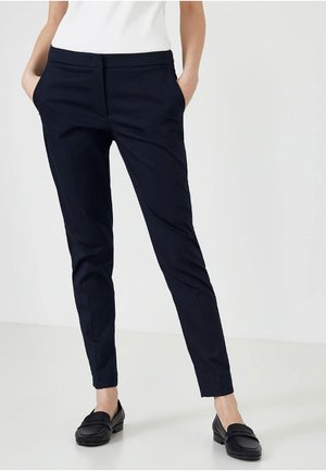Vrouw die slanke donkerblauwe broek met zijzakken draagt, gecombineerd met zwarte loafers en een wit topje, staand tegen een eenvoudige lichte achtergrond.