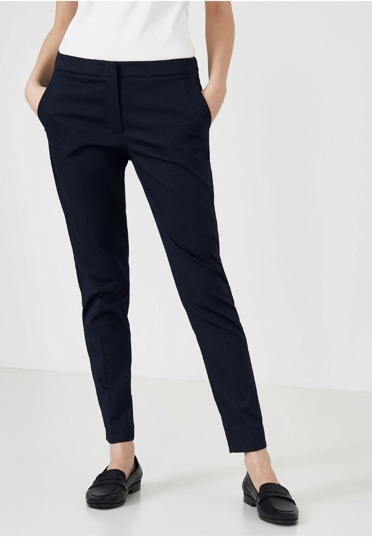 Femme portant un pantalon slim bleu marine foncé avec poches latérales, associé à des mocassins noirs et un haut blanc, debout devant un fond clair uni.