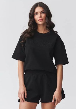 Młoda kobieta z długimi kręconymi włosami, ubrana w czarny oversize'owy t-shirt i pasujące czarne spodenki, stoi na jednolitym jasnym tle.