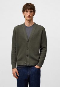 Ung man som bär en mörkgrön knappad cardigan över en grå t-shirt och blå jeans, står med en hand i fickan mot en enfärgad bakgrund.