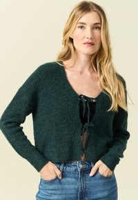 Maglione corto verde scuro realizzato in morbido tessuto a maglia, caratterizzato da una profonda scollatura a V e chiusura con lacci regolabile nella parte anteriore.