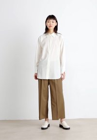 Witte langwerpige blouse met een puntige kraag, gecombineerd met olijfgroene broek met wijde pijpen en witte instapschoenen met zwarte zolen.