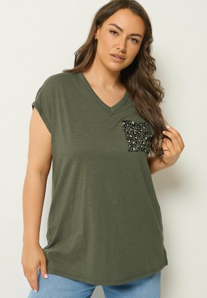 Vrouw met lang bruin haar die een olijfgroen T-shirt met V-hals en een luipaardprintzak draagt, poseert tegen een lichte achtergrond.