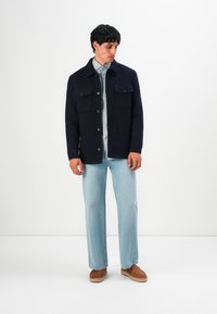 Mann in einer dunkelblauen knöpfbaren Jacke, hellblauem Hemd, hellblauen Jeans und braunen Slippern, der vor einem weißen Hintergrund steht.