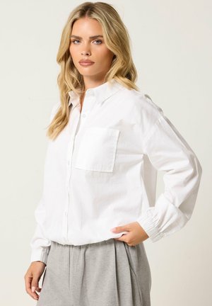 Femme blonde portant une chemise blanche boutonnée avec une poche poitrine et un pantalon gris plissé, posant avec une main dans la poche.