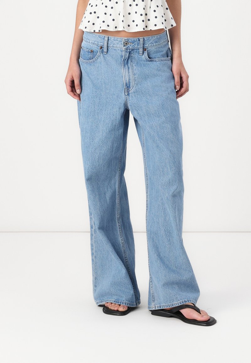 Vaqueros de mezclilla de corte acampanado en azul claro con cintura alta, diseño de cinco bolsillos y textura desgastada, combinados con chanclas negras.