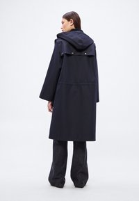 Αδιάβροχο παλτό σε σκούρο μπλε με κουκούλα, που διαθέτει πίσω πτερύγιο και ζώνη στη μέση. Μακριά μανίκια και ίσια γραμμή, κατάλληλο για στρώσεις ρουχισμού.