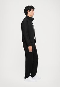 Zwarte sportjack met een hoge kraag, gecombineerd met zwarte broek en witte sneakers. Glad materiaal en relaxed fit. Zijprofielweergave.