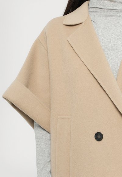 Beige wool-blend coat with short sleeves, notched collar, double button closure, and side pockets; layered over a gray turtleneck.